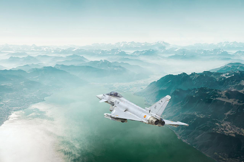 A Eurofighter jet is flying over a scenic, cloudy mountain range.