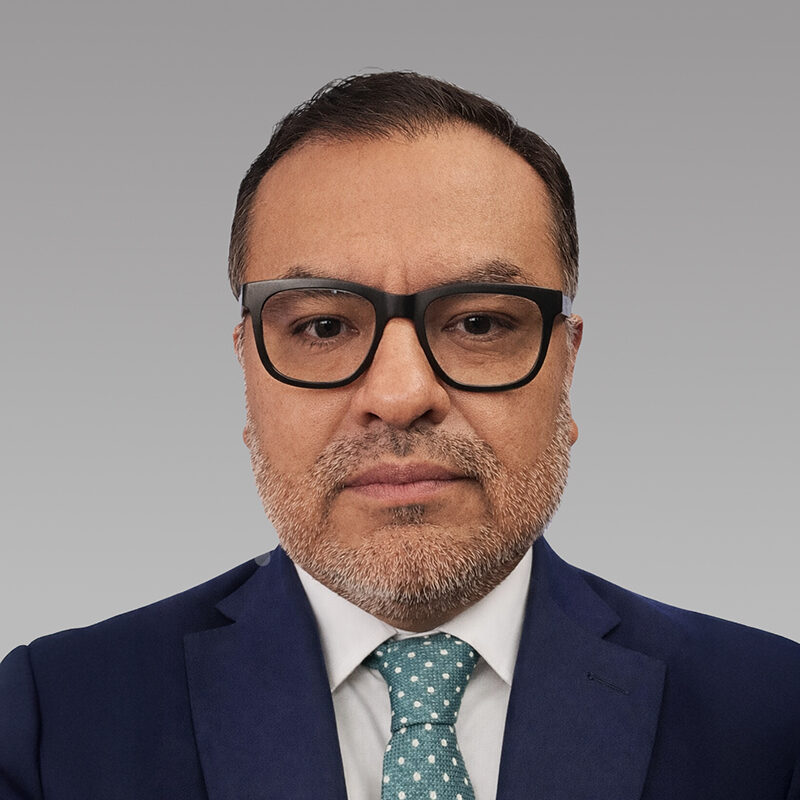 Omar Elias is a distinguished man with large black glasses. He stares into the camera with a serious expression.